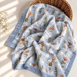 A light blue muslin baby blanket with embroidered woodland characters and small white flowers, draped over a woven basket on a sunlit cream surface with soft shadows and small white flowers nearby.
