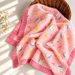 A pink muslin baby blanket with cartoon cat prints and yellow stars, draped over a brown wicker basket on a sunlit white surface with soft shadows.