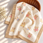 A soft cotton muslin baby blanket with a teddy bear print, draped over a brown wicker basket on a white surface with soft sunlight shadows.