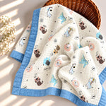 A quilted baby blanket with a light blue border and an off-white center printed with cute cartoon animals, displayed in a wicker basket with a small flower arrangement on a sunlit cream surface.