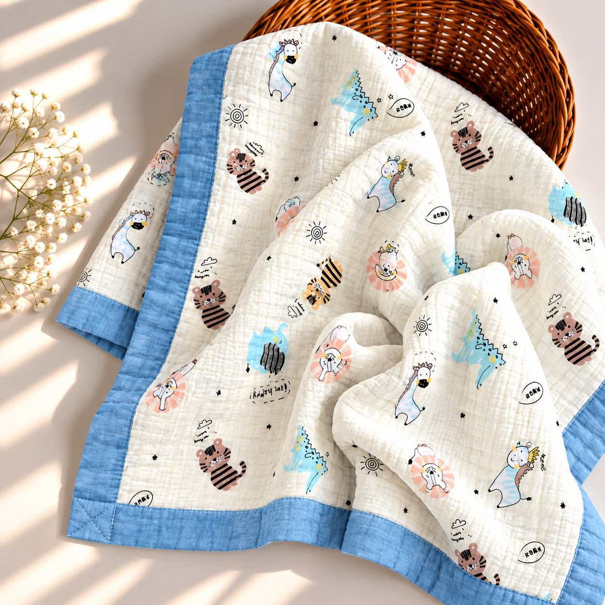 A quilted baby blanket with a light blue border and an off-white center printed with cute cartoon animals, displayed in a wicker basket with a small flower arrangement on a sunlit cream surface.