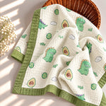 A quilted baby blanket with a green dinosaur and avocado print, draped over a woven basket on a sunlit surface.